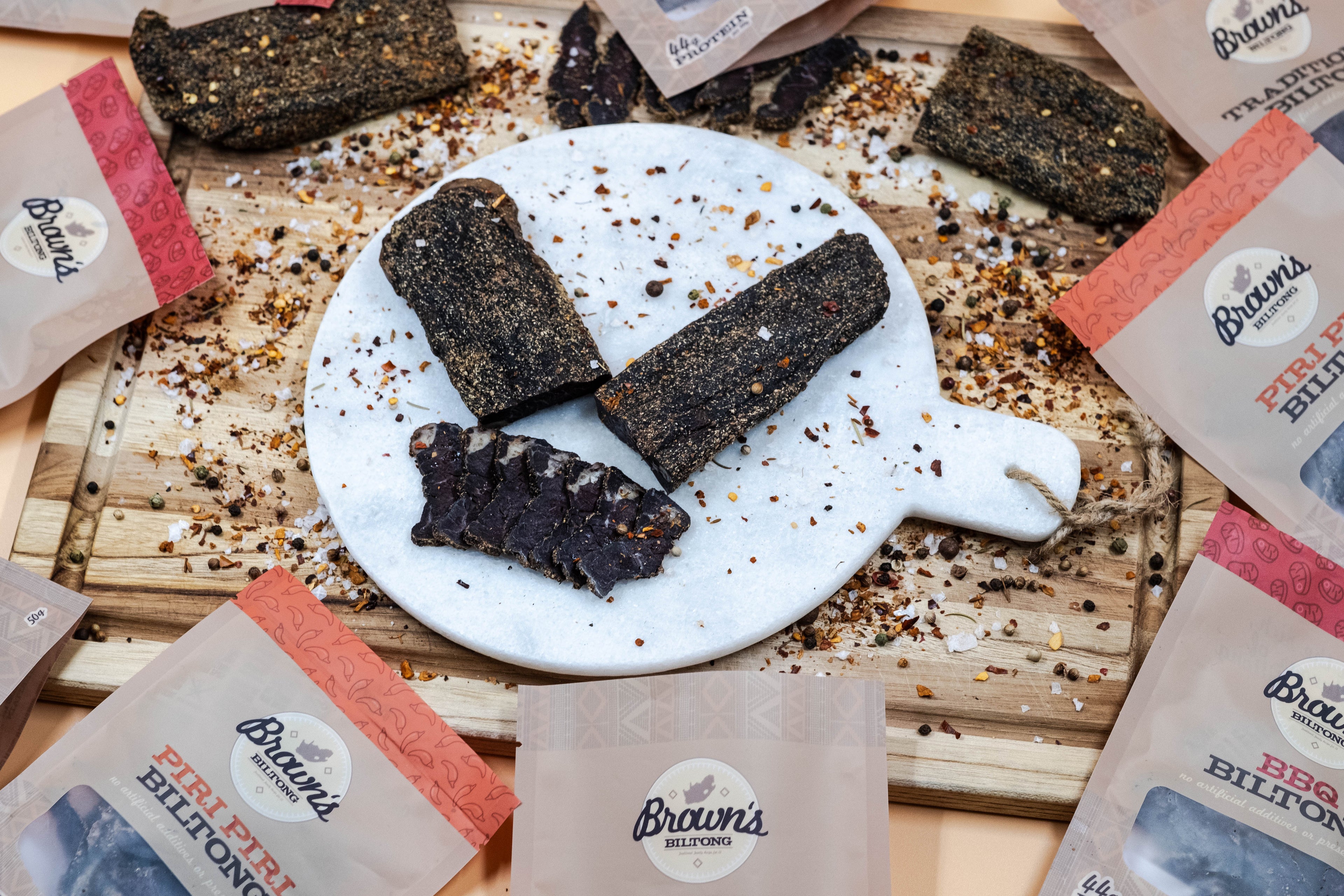 Sliced biltong on a white granite chopping board has packets of biltong scattered around the outside.