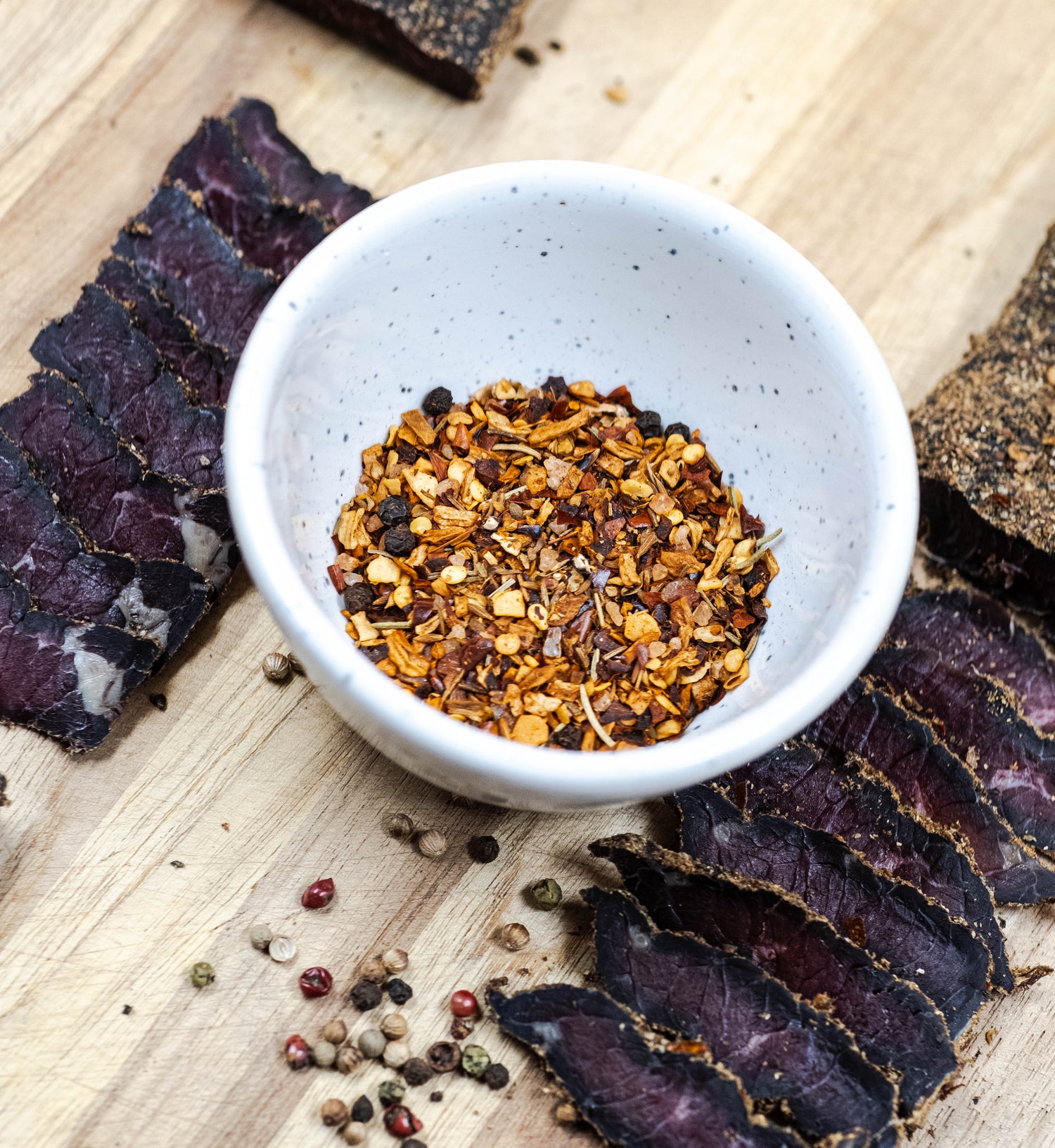 Sliced biltong on an oak chopping board, spices have been combined in a small porcelain bowl and some peppercorns are scattered across the chopping board. 