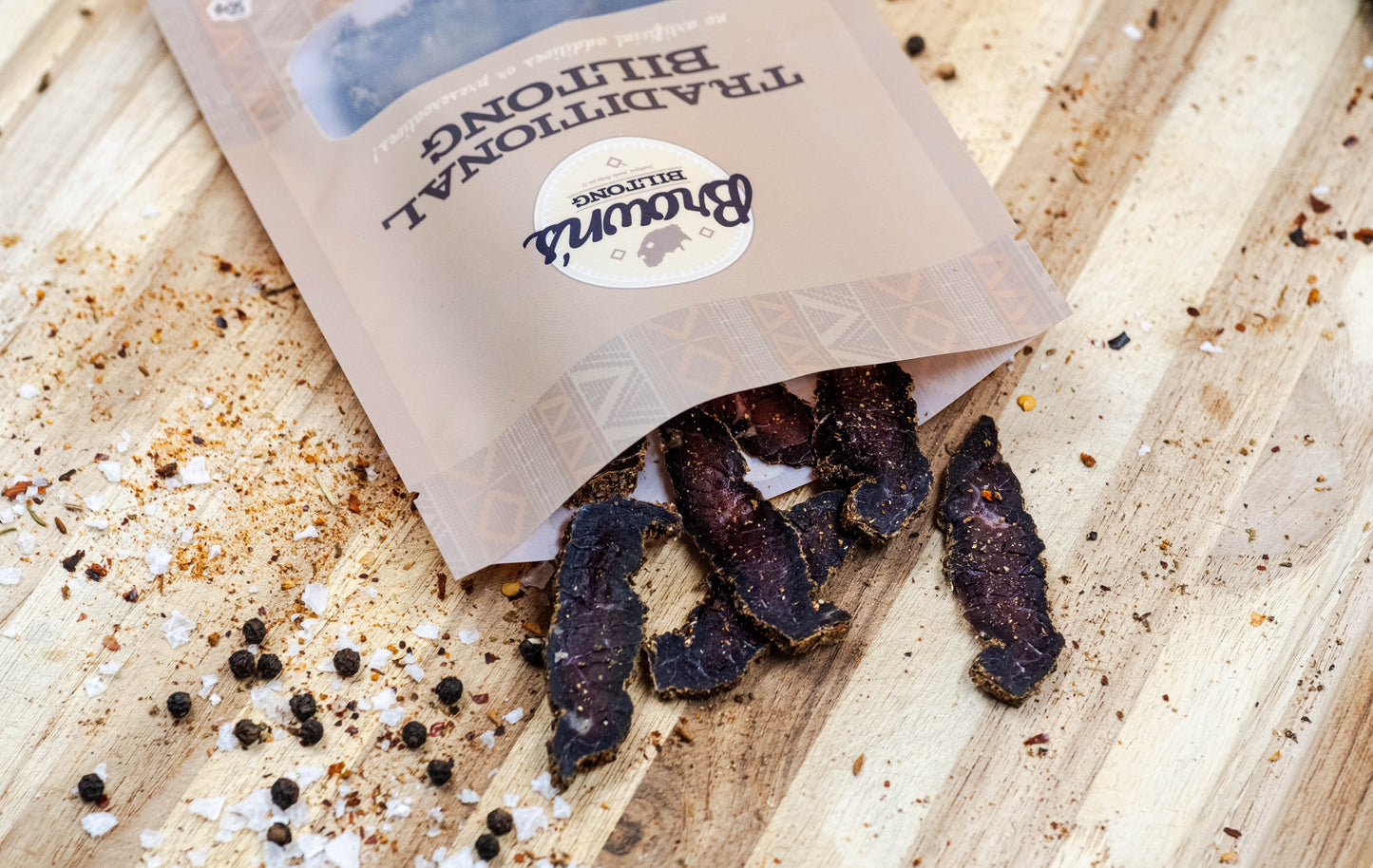 A single pack of traditional flavour biltong is laid on an oak chopping board. The packet is open and some pieces have fallen out onto the chopping board and are surrounded by a mixture of spices