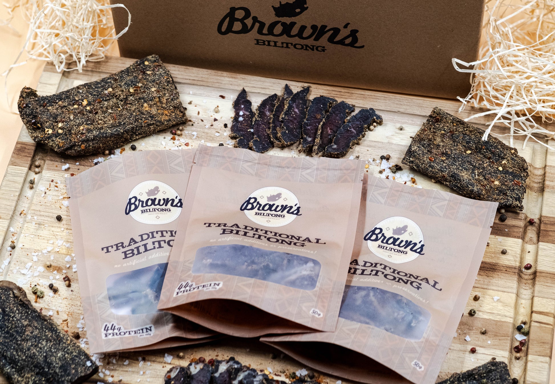 3 packets of traditional biltong are laid on an oak chopping board with whole steaks of biltong scattered around them along with some spices